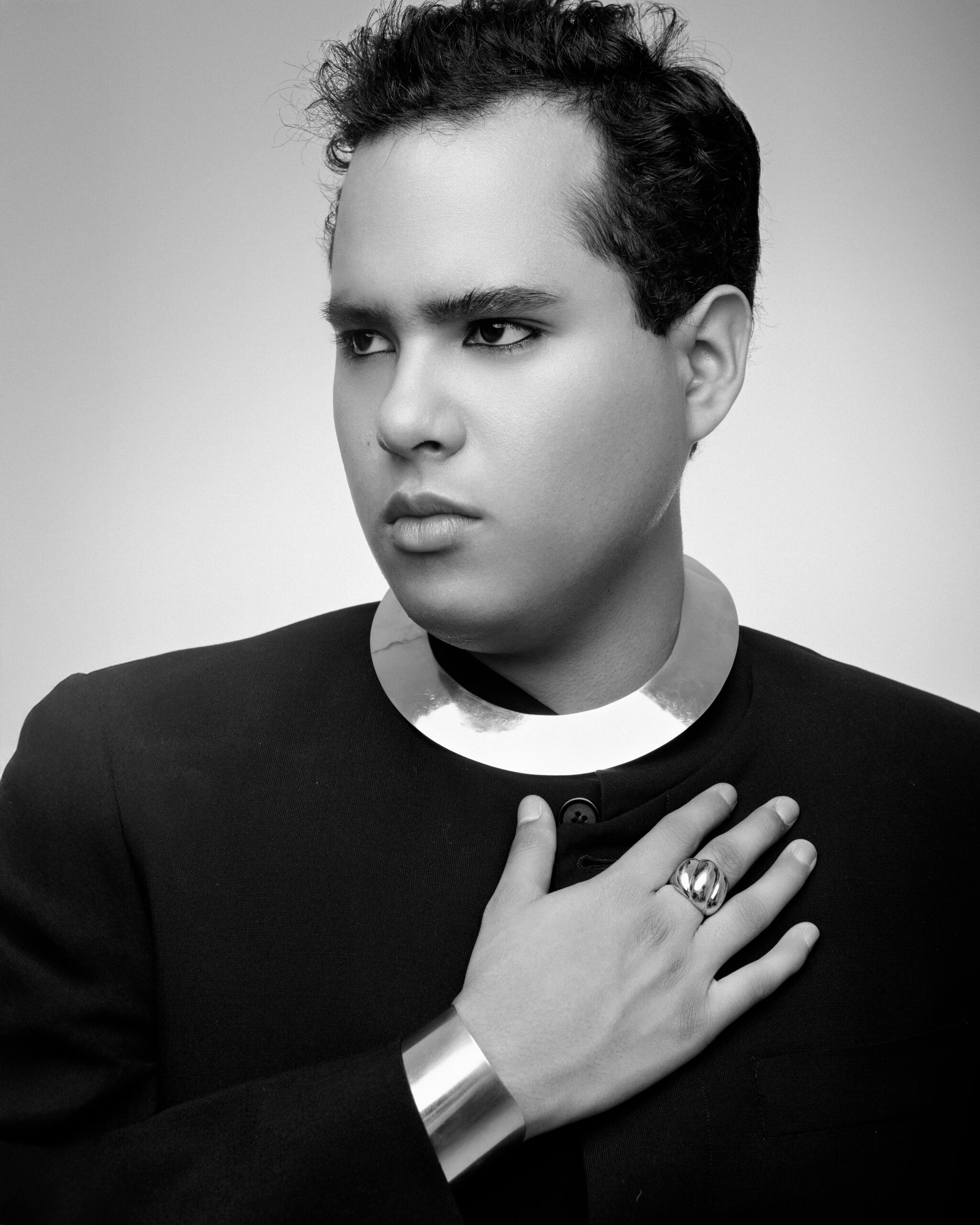 Portrait of a person with a metallic collar and bracelet, wearing a black outfit and striking a serious pose.