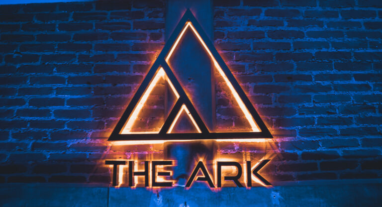 Señal neón de The Ark Club Roma con luz dorada y neón azul sobre muro de ladrillo, marcando el ritmo del sintetizador.