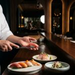 A sushi chef serves fresh omakase nigiri beside sashimi plates on a polished wooden counter in a dim, elegant Japanese dining room in Roma Condesa.