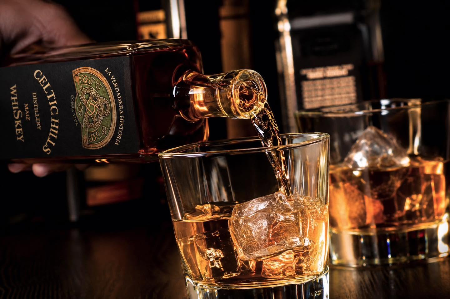 Celtic Hills whiskey pours over ice into a glass, with amber reflections and a dark bar backdrop for the 100% Mexican maize spirit in a moody setting.