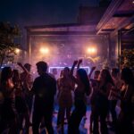 En The Ark Club Roma, un grupo baila mientras un DJ mezcla música electrónica bajo luces violetas y cálidas de verano.